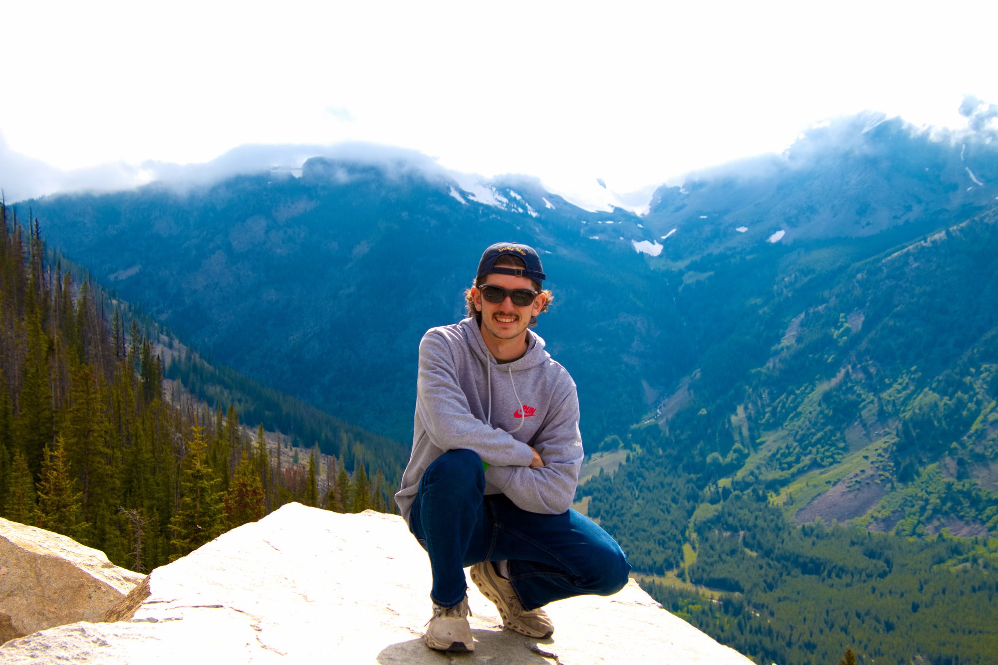 Donald Voetberg crouched on a rock outcrop with forested mountains and snow-capped peaks behind him